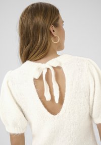 Top tricoté blanc avec des manches courtes bouffantes et un dos ouvert, présentant une fermeture à nœud. Texture douce et apparence chaleureuse.