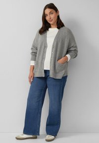 Cardigan grigio a maglia con vestibilità rilassata, caratterizzato da un colletto a V, bottoni e polsini a costine, abbinato a jeans blu a gamba larga e sneakers.