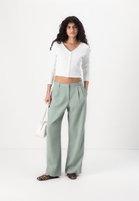 Gilet court à côtes blanches, pantalons larges vert clair et ballerines à imprimé léopard. Modèle tenant un sac à main blanc. Design simple et moderne.