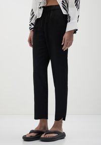 Personne portant un pantalon noir ample avec des fentes aux chevilles, des sandales noires, un bracelet de cheville en chaîne dorée, et une chemise blanche avec des motifs de feuilles noires.