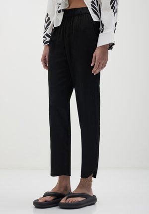 Persona che indossa pantaloni neri larghi con spacchi alle caviglie, sandali neri, un braccialetto d'oro alla caviglia e una camicia bianca con motivi foglia neri.