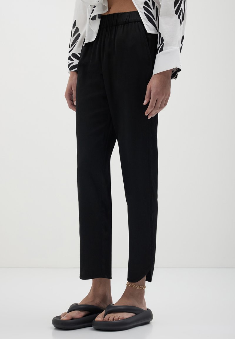 Personne portant un pantalon noir ample avec des fentes aux chevilles, des sandales noires, un bracelet de cheville en chaîne dorée, et une chemise blanche avec des motifs de feuilles noires.