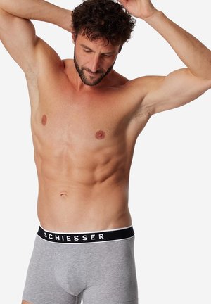 Oberkörperfreier Mann, der die Arme über dem Kopf anpasst, trägt graue Boxershorts mit schwarzem Bund mit der Aufschrift "SCHIESSER" vor einfachem weißem Hintergrund.