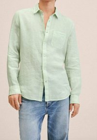 Chemise en lin vert clair avec col boutonné, poche plaquée et manches longues, associée à un jean en denim bleu.