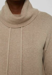 Pull à col roulé beige en tissu tricoté côtelé, avec un col plié et deux coutures verticales sur le devant.
