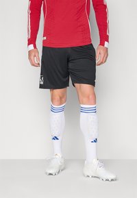 Maillot à manches longues rouge avec des accents blancs, shorts noirs, chaussettes blanches avec des rayures bleues, et crampons blancs. Logo sportif sur le short.