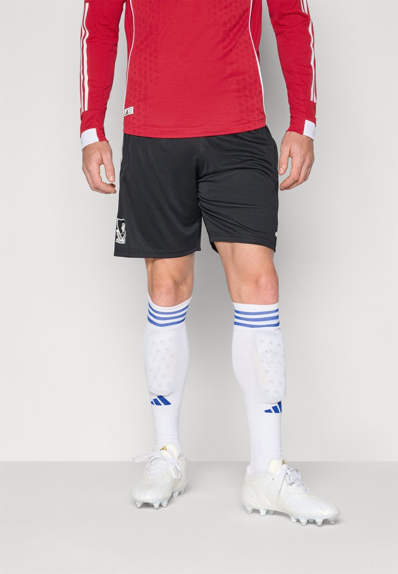 Maillot à manches longues rouge avec des accents blancs, shorts noirs, chaussettes blanches avec des rayures bleues, et crampons blancs. Logo sportif sur le short.