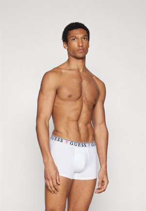 Boxer da uomo bianchi con elastico nero che presenta il logo "GUESS". Modello in piedi con una posa neutra, mostrano il fit e il design del capo.