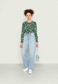 Blusa longa de mangas compridas verde com flores e ombros franzidos, acompanhada de jeans de cintura alta azul claro; completando o look com ténis brancos e uma bolsa azul.