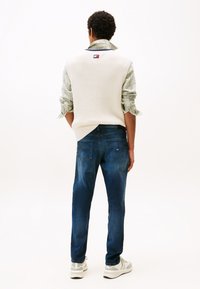 Biały dziergany bezrękawnik na zielonej koszuli w kratę, zestawiony z ciemnoniebieskimi dżinsami i szarymi trampkami. Minimalistyczny design, luźny krój.