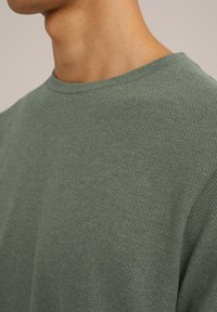 Zelen steksturiran pulover s okroglenim izrezom, izdelan iz tkane tkanine, s tesno prileganjem in mehkobo ter rahlo puhaste površine.