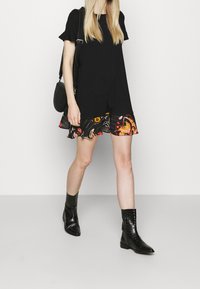 Czarna sukienka t-shirtowa z rozkloszowanym dołem w kwiatowy wzór, zestawiona z czarnymi botkami za kostkę. Modelka niesie małą czarną torebkę na ramię.