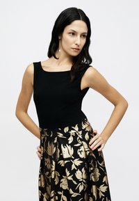 Μαύρο εφαρμοστό τοπ με τετράγωνη λαιμόκοψη, συνδυασμένο με μαύρη σατέν φούστα που ρέει και έχει μεταλλικά χρυσά λουλουδάτα σχέδια.