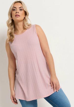 Blonde Frau, die ein ärmelloses, geripptes rosa Tunika-Oberteil und eine blaue Jeans trägt, steht vor einem schlichten hellen Hintergrund.