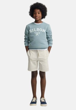 Niño con cabello rizado que lleva una sudadera azul clara con el texto "SILBON", pantalones cortos beige y zapatillas azules, de pie con las manos en los bolsillos.