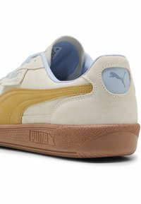 Puma-tennis chaussées en daim beige avec une rayure jaune, des accents bleu clair et une semelle en caoutchouc brun texturé. Présente un logo sur le talon.