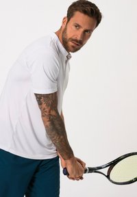 Polo shirt bianco a maniche corte con finitura testurizzata, abbinato a pantaloni corti blu scuro. Una persona tiene una racchetta da tennis con un'impugnatura decorata.
