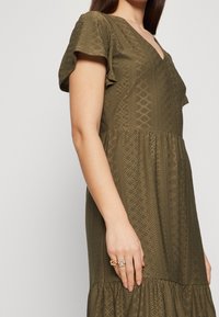 Oliivinvihreä mekko, jossa on lyhyet röyhelöhihat, V-pääntie, timantinmuotoista kuvioitua tekstuurikangasta ja liehuva helma. Kädet koristeltu sormuksilla.