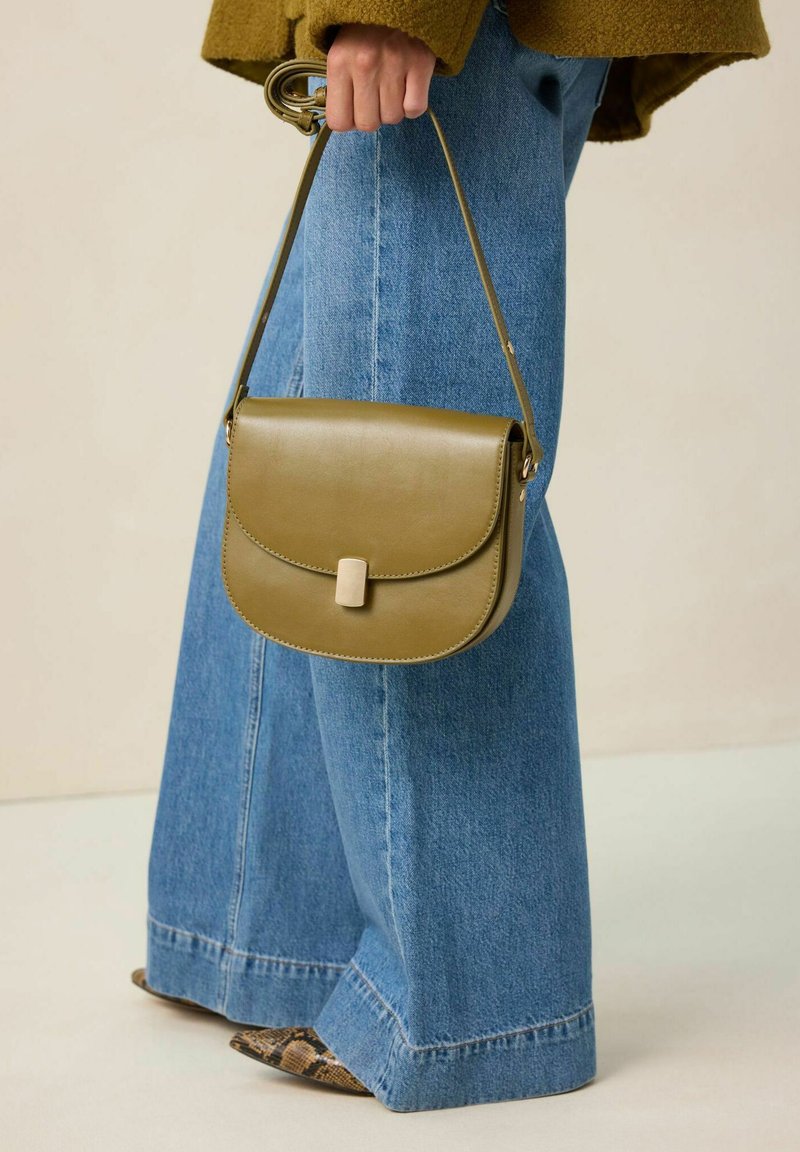 Sac en cuir vert olive avec un rabat arrondi, un fermoir en métal et une bandoulière. Associé à un jean en denim bleu à jambes larges et des chaussures à motifs.