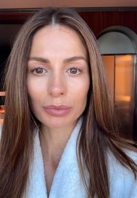 Kvinna med långt, rakt brunt hår som bär en vit morgonrock. Naturlig make-up med fylliga läppar och mjuka drag. Bakgrunden visar en suddig interiör.