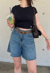 Camiseta negra ajustada, pantalones cortos de mezclilla azules, cinturón de cuero marrón con hebilla de plata. Vaso transparente con bebida verde. Bolso bandolera marrón.