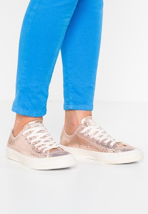Personne portant un pantalon bleu vif et des baskets métalliques effet peau de serpent couleur or rose avec des lacets et semelles blanches sur un sol blanc.