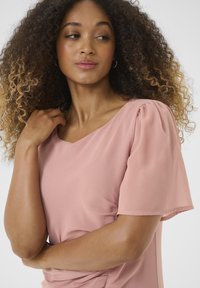 Pink short-sleeve blouse with a v-neck, featuring soft, flowing fabric and gentle pleating at the shoulders. Model poses with hand on chest.