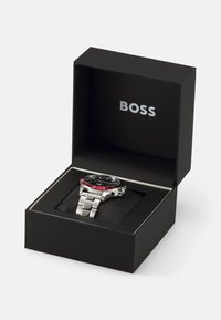 Montre BOSS dans une boîte noire. Dotée d'un bracelet en métal argenté, d'un cadran noir, d'un accent rouge sur la lunette, et d'un affichage de la date à 3 heures.