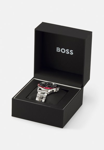 Montre BOSS dans une boîte noire. Dotée d'un bracelet en métal argenté, d'un cadran noir, d'un accent rouge sur la lunette, et d'un affichage de la date à 3 heures.