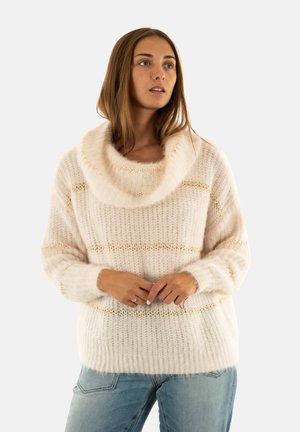 Donna con indosso un maglione spesso color crema con un ampio collo a cascata e strisce orizzontali dorate, abbinato a jeans blu chiaro.