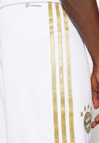 Vita idrottsshorts med guldstripes längs sidorna, med FC Bayern Münchens logotyp och "AEROREADY"-märkning vid midjan.