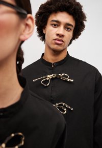 Juodas marškinis su stovinčia apykakle, puoštas raštuotais audinio raiščiais. Artimas modelio veido ir profilio kadras su minkštu fokusu fone.