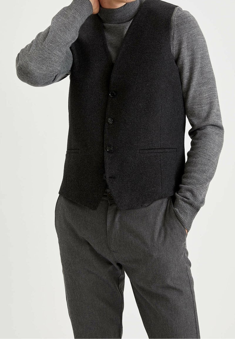 Schwarze Wollweste mit V-Ausschnitt, ausgestattet mit sechs Knöpfen und zwei Fronttaschen, die über einem grauen Strickpullover und grauen Hosen getragen wird.