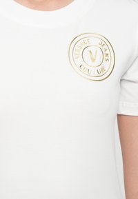 T-shirt branca de algodão com um decote redondo, apresentando um logótipo em relevo dourado da Versace Jeans Couture e elementos circulares no peito.