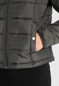 Mørkegrøn pufferjakke med lynlås, horisontale syninger, sidelomme og sølvfald. Glat, skinnende tekstur.