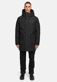 Melna parka ar rāvējslēdzēja kabatu un pogu aizdari, ar kapuci, divām priekšējām kabatām un taisnu siluetu. Valkāta ar melnām biksēm un zābakiem.