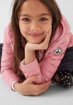 Jeune fille souriante aux longs cheveux bruns, appuyant son menton sur sa main, portant une veste rose avec un écusson rond et un pantalon foncé avec des chaussures cloutées.