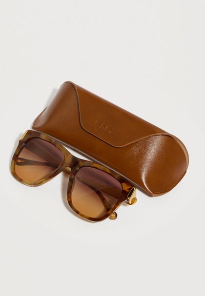 Γυαλιά ηλίου με καφέ φακούς διαβαθμισμένης απόχρωσης σε σκελετό τύπου καβούκι δίπλα σε καφέ δερμάτινη θήκη προστασίας πάνω σε λευκή επιφάνεια.