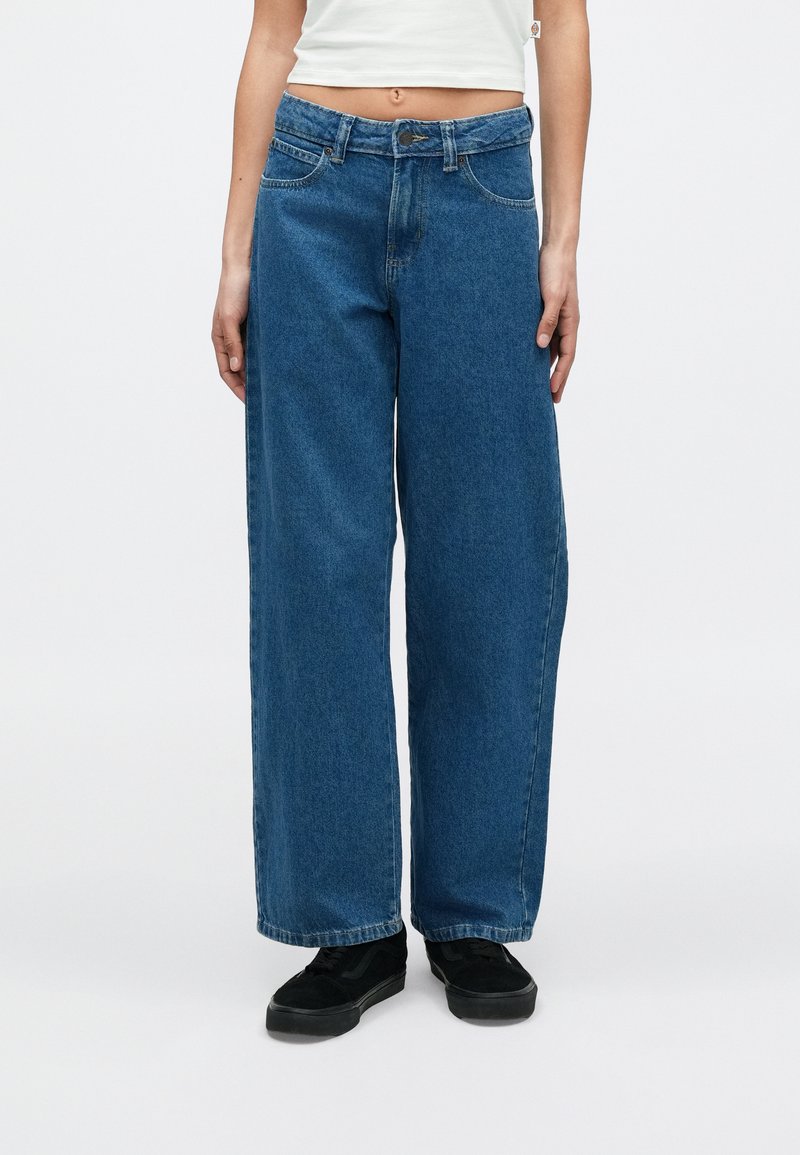 Personne portant un jean bleu taille haute à jambes larges avec des baskets noires et un top blanc court, debout devant un fond blanc uni.