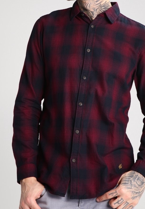 Homme portant une chemise à carreaux rouge et noire à boutons, avec des mains et un cou tatoués, debout devant un fond clair uni.