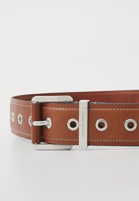 Ruskea nahkavyö, jossa on hopeanvärisiä yksityiskohtia, suuret niitit ja erottuva valkoinen ompelu, brändätty tekstillä "MICHAEL KORS."