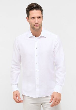 Mand iført en tætsiddende hvid skjorte med knapper og lange ærmer, stående mod en ensfarvet hvid baggrund, kiggende let til siden.