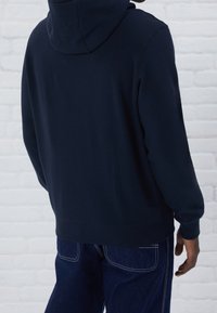 Felpa navy con cappuccio regolabile, polsini e orlo a costine. Design aderente con texture liscia, indossata con pantaloni in denim blu con cuciture bianche.