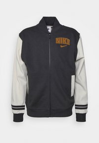 Nike zip-up jack met donkergrijs en crèmekleurige tinten, geribbelde manchetten, zijzakken en het Nike-logo in oranje op de borst.