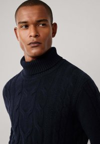 Muž v tmavě námořnickém svetru s kabelovým pletením a vysokým límcem se dívá na stranu na pozadí s jednoduchým světlým pozadím.