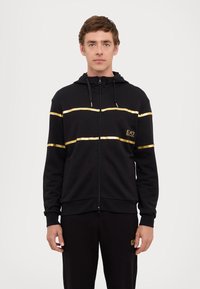 Black zip-up hoodie with gold striped accents and logo. Made of soft material, features a hood and drawstrings, ribbed cuffs and hem.