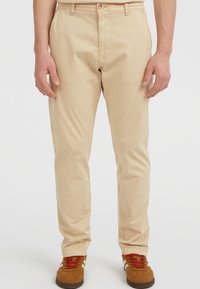 Man in beige chinos en brown sneakers met rode veters, staande tegen een effen witte achtergrond.