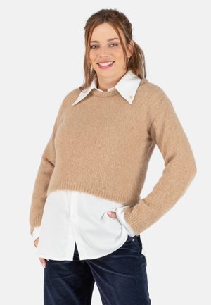 Pull court marron clair en matière douce, porté sur une chemise blanche à col. Il a des manches longues et un ourlet côtelé.