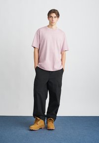 Tricou roz deschis, supradimensionat, cu un logo mic, asortat cu pantaloni cargo negri, lejeri, și botine cu șireturi bej, pe un covor albastru.