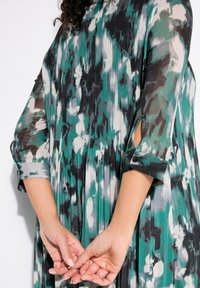 Robe plissée à motifs floraux dans des nuances de vert, noir et blanc. Dotée de manches 3/4 avec des poignets et de découpes sur les côtés. Tissu léger.